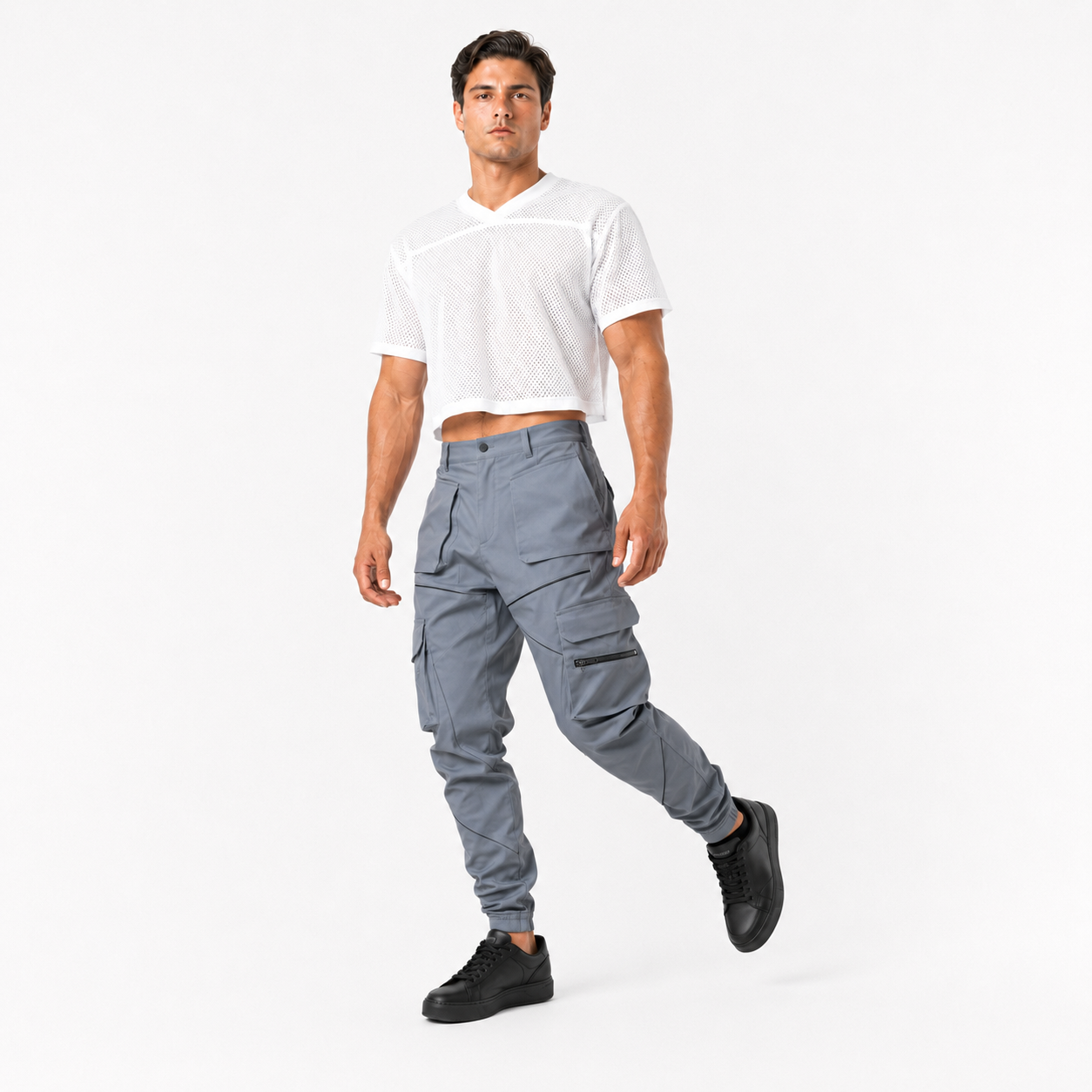 A male model wearing a white cropped V-neck t-shirt and slate gray cargo pants with black sneakers, walking against a plain white studio background.